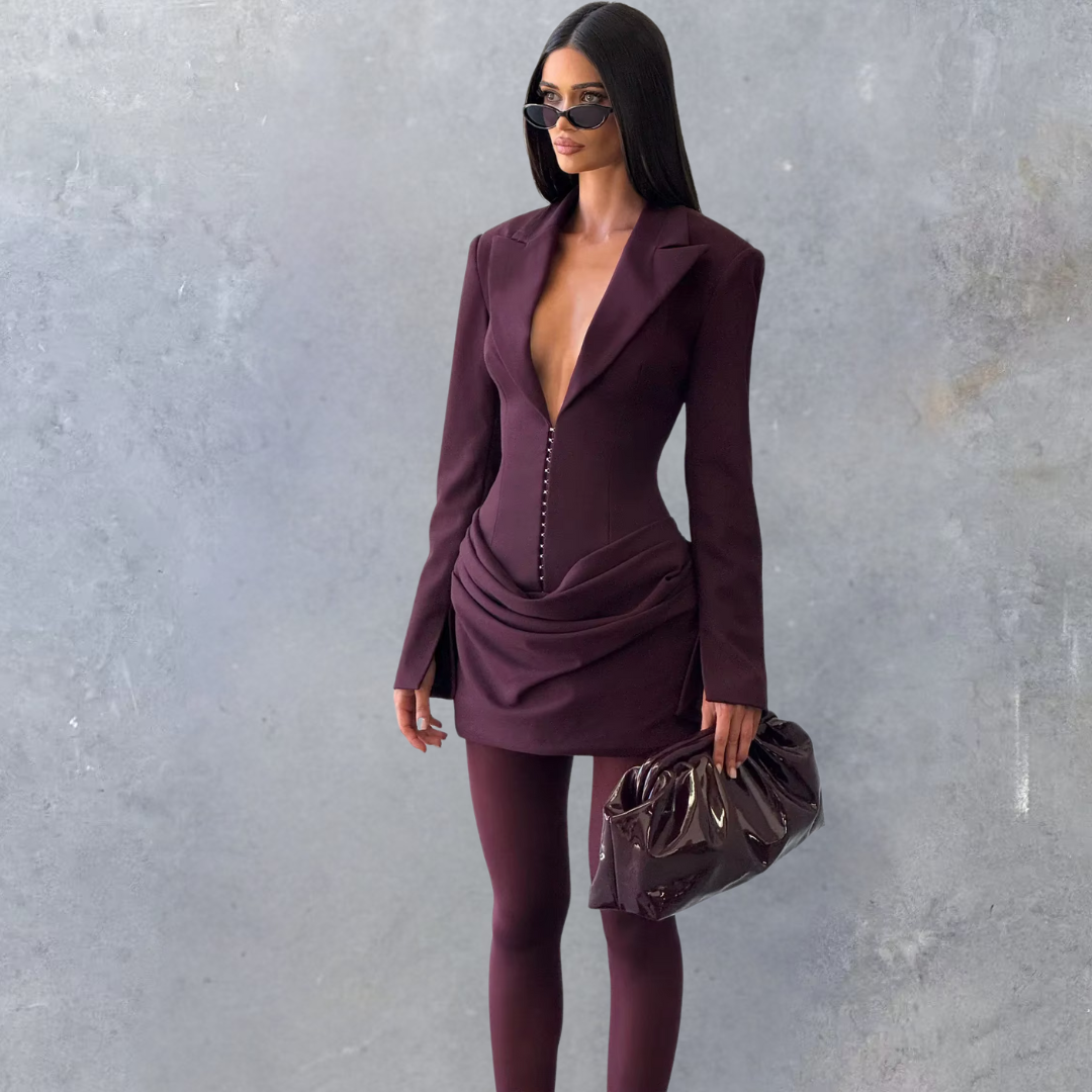 Woman in a deep purple outfit with sunglasses and a handbag against a gray background