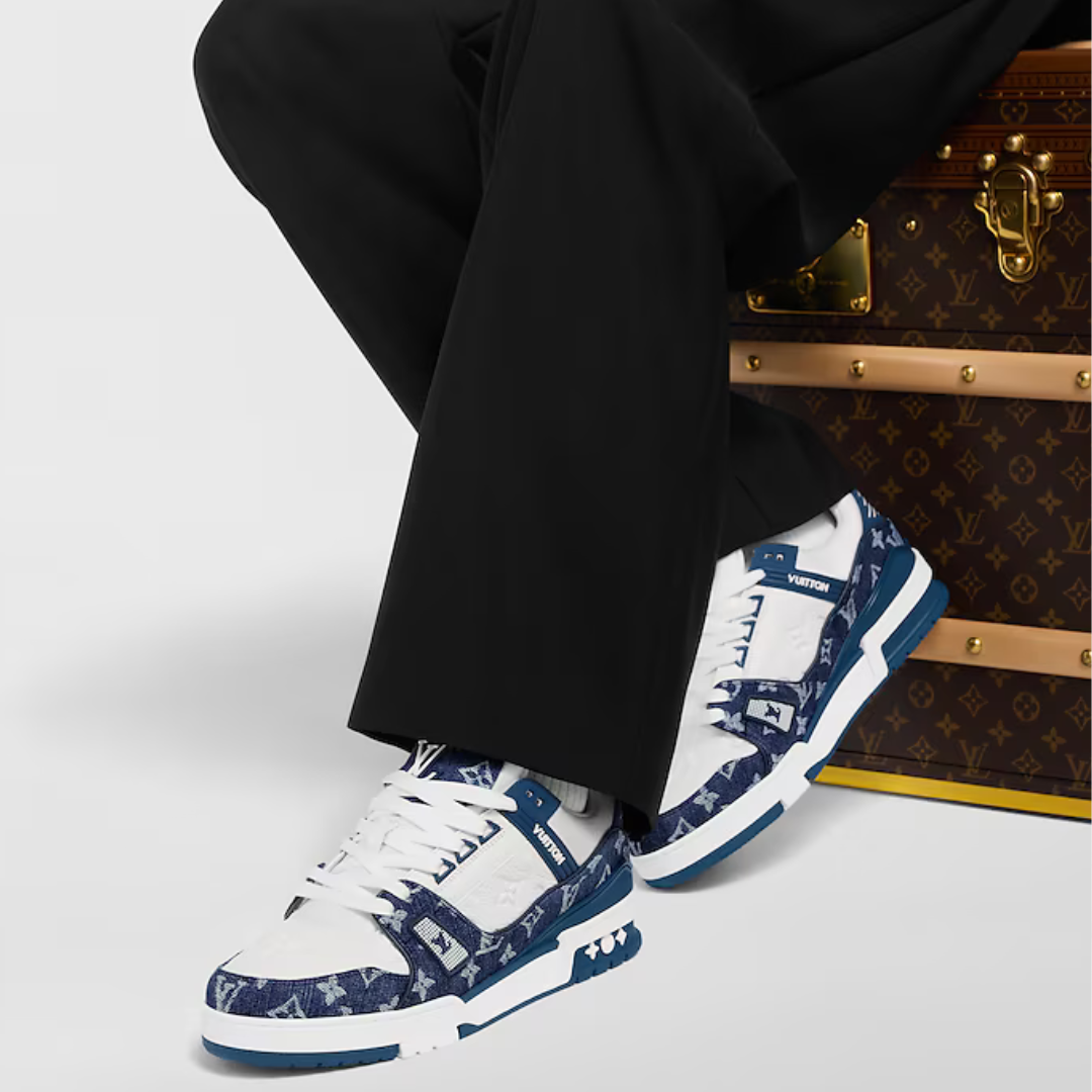 White sneakers with blue pattern worn by a person sitting next to a Louis Vuitton suitcase.